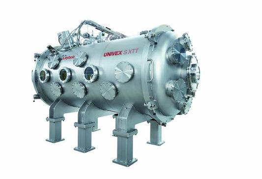 A large, metallic, cylindrical Space Simulation System standing on four sturdy legs. The device features multiple circular ports and panels labeled with "Leybold North America Univex SXT". Various pipes and cables are connected at the top. The overall color is silver.